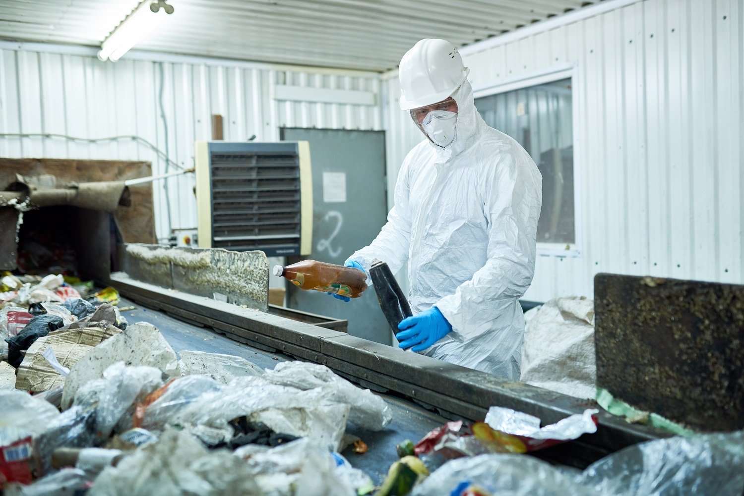Portrait of worker wearing biohazard suit and hardhat working at waste processing plant sorting recyclable materials on conveyor belt